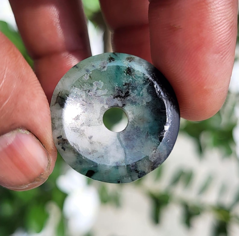 May include: A circular, flat pendant with a central hole, featuring a polished surface with a mix of translucent green, white, and dark grey inclusions. The stone displays natural mineral patterns and a smooth, reflective finish throughout.