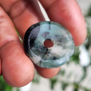 May include: A circular, donut-shaped pendant made of polished stone, featuring a marbled pattern of deep forest green, translucent light green, and opaque white, with dark black inclusions and a smooth, glossy finish throughout.