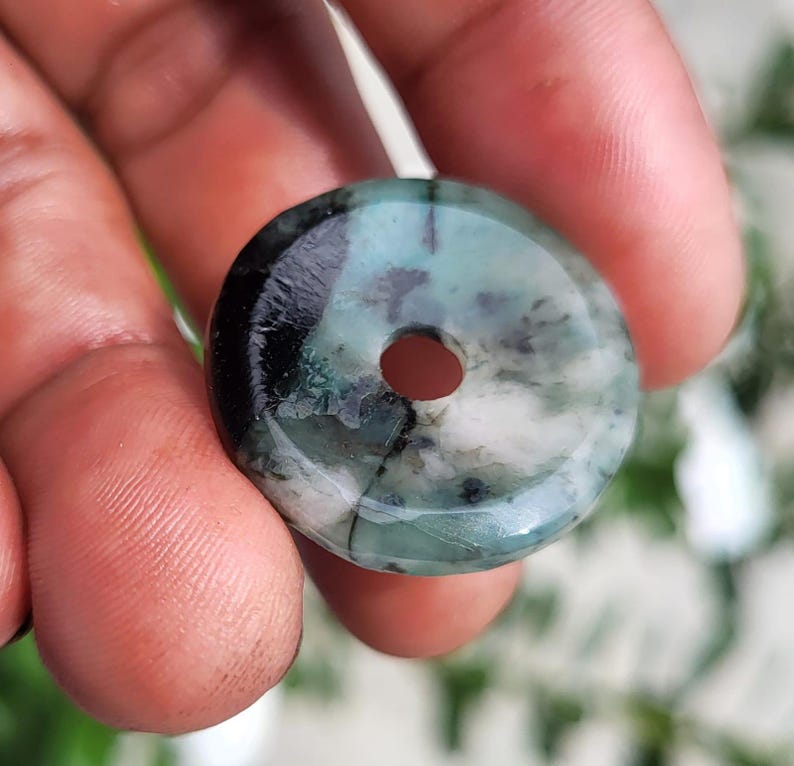 May include: A circular, donut-shaped pendant made of polished stone, featuring a marbled pattern of deep forest green, translucent light green, and opaque white, with dark black inclusions and a smooth, glossy finish throughout.