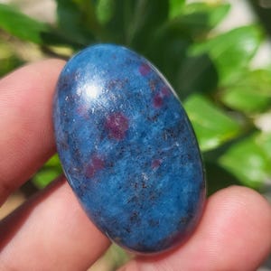May include: An oval-shaped cabochon gemstone featuring a deep blue base colour with scattered, distinct magenta-red inclusions and dark, fine mineral flecks throughout the polished, smooth surface of the stone, reflecting light on its top.