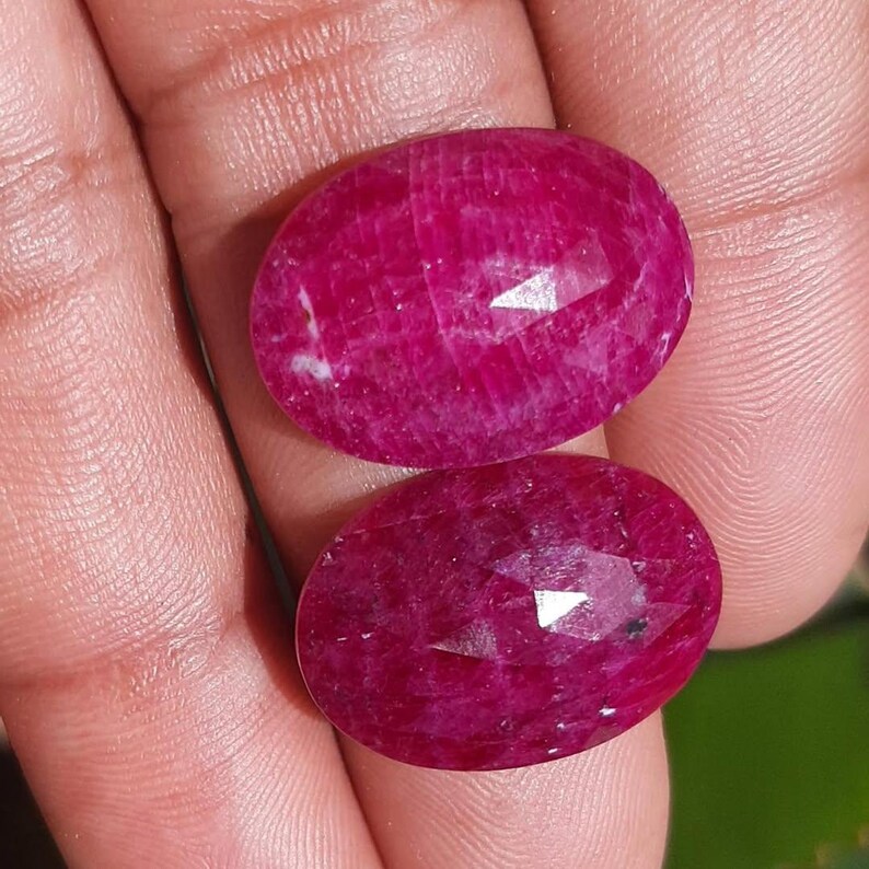 May include: Two oval-shaped, faceted ruby gemstones. The rubies are a deep, vibrant red color with a polished surface. The gemstones are held in a hand, showcasing their size and brilliance. The facets catch the light, creating a sparkling effect.