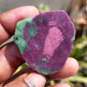 May include: A close-up of a natural gemstone, showcasing a striking colour contrast. The stone has a deep magenta core, a lighter pink ring, and a green outer edge. The stone is held in a hand.