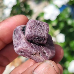 Könnte beinhalten: Ein rohes, unpoliertes Rubin-Kristallcluster mit einem tiefen Magenta- und Violettton sowie sichtbaren schwarzen Mineraleinschlüssen. Der Stein hat eine raue, texturierte Oberfläche mit natürlichen geometrischen Facetten.