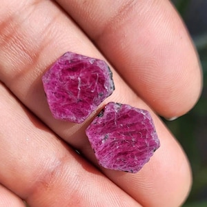 May include: Two rough-cut, hexagonal ruby gemstones. The rubies are a deep, vibrant pinkish-red colour with visible internal structures and dark inclusions. The gemstones are displayed against a blurred background.