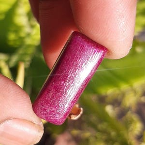 May include: A rectangular, polished gemstone in a deep magenta hue. The stone is held between fingers, showcasing its smooth, reflective surface and subtle internal patterns. The background is blurred, with green foliage.