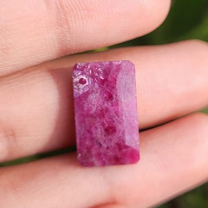 May include: A rectangular, faceted, deep pink ruby gemstone. The gemstone has a natural, rough texture and a hexagonal pattern on one end. The ruby is held in a hand, with a blurred green background.