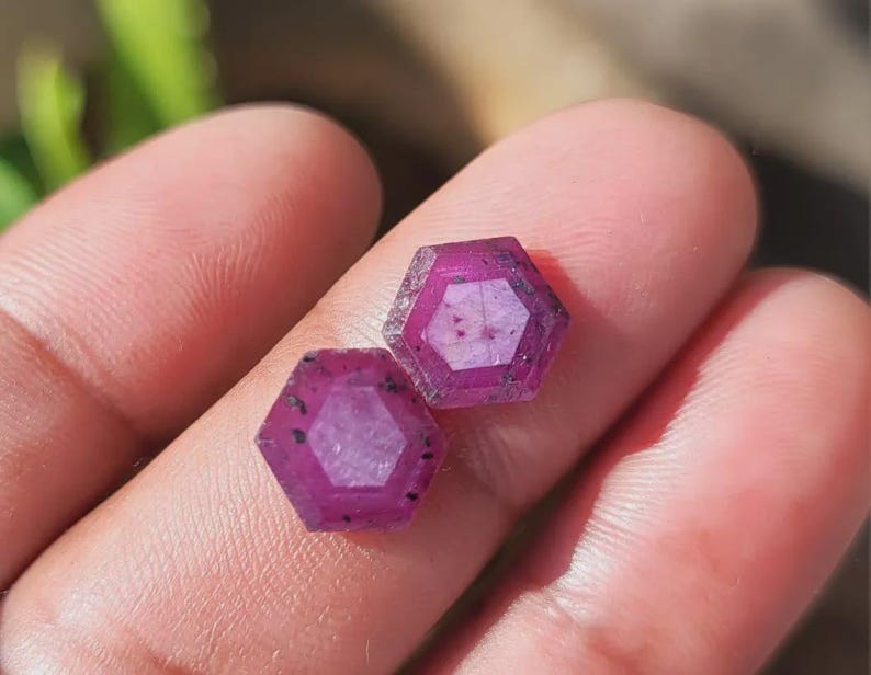 May include: Two hexagonal, faceted ruby gemstones. The rubies are a deep pinkish-red color with visible internal inclusions. The gems are displayed on a light-toned background, with a blurred green plant in the background.