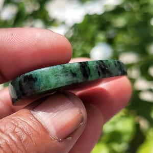 May include: A polished, curved band of green stone featuring prominent black inclusions throughout its surface. The material has a smooth, glossy finish with a translucent quality, showcasing a natural mottled pattern of deep forest green and dark charcoal.