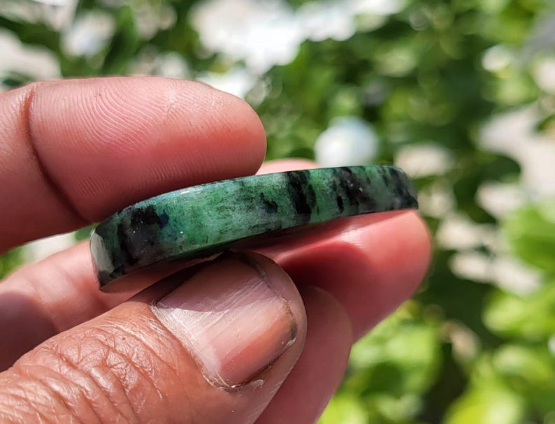 May include: A polished, curved band of green stone featuring prominent black inclusions throughout its surface. The material has a smooth, glossy finish with a translucent quality, showcasing a natural mottled pattern of deep forest green and dark charcoal.