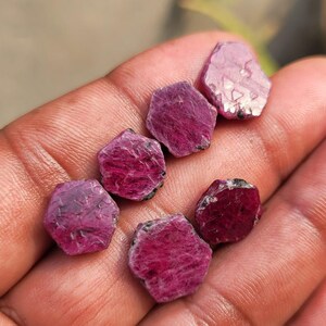 May include: A collection of six rough-cut ruby gemstones, each with a hexagonal shape and a deep red-pink hue. The gemstones have a natural, unpolished appearance, with visible textures and variations in color. The rubies are held in a hand, showcasing their size and color.
