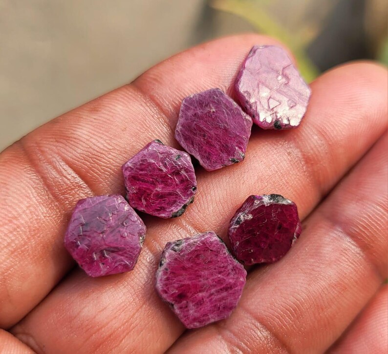 May include: A collection of six rough-cut ruby gemstones, each with a hexagonal shape and a deep red-pink hue. The gemstones have a natural, unpolished appearance, with visible textures and variations in color. The rubies are held in a hand, showcasing their size and color.