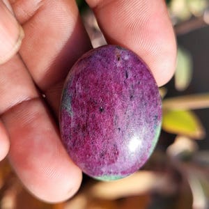 Puede incluir: Una gema ovalada de color magenta intenso, salpicada de manchas oscuras y toques de verde. La superficie pulida refleja la luz, mostrando la textura natural y las variaciones de color de la piedra. Sostenida en una mano, la gema es un ejemplo llamativo de belleza natural.