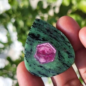 May include: A teardrop-shaped polished gemstone featuring a vibrant magenta hexagonal crystal inclusion centred within a mottled green and black zoisite matrix. The stone has a smooth, flat surface with visible natural mineral patterns throughout.