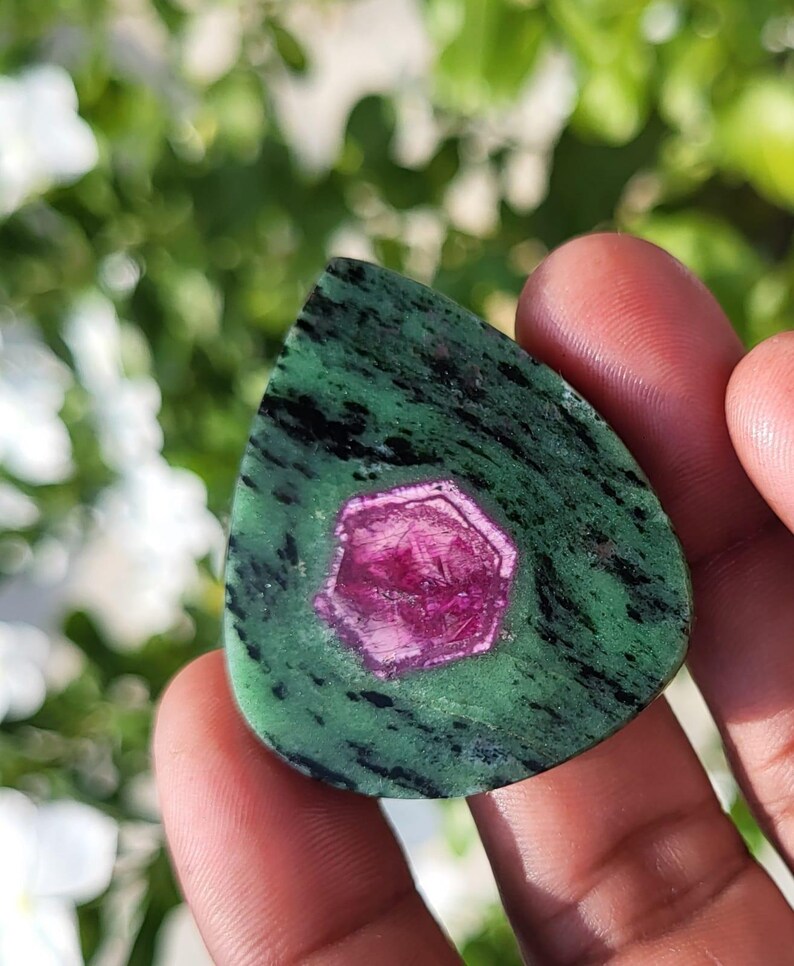 May include: A teardrop-shaped polished gemstone featuring a vibrant magenta hexagonal crystal inclusion centred within a mottled green and black zoisite matrix. The stone has a smooth, flat surface with visible natural mineral patterns throughout.