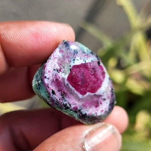 Könnte beinhalten: Nahaufnahme eines Edelsteins, der in einer Hand gehalten wird. Der Stein hat einen leuchtenden, tiefroten Mittelpunkt, umgeben von Weiß-, Rosa- und Grüntönen. Die Oberfläche des Steins hat eine leicht raue Textur.