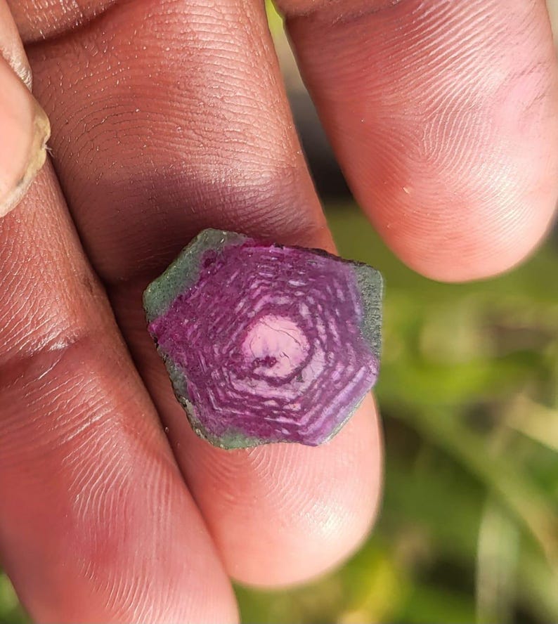 Pode incluir: Uma gema de turmalina melancia cortada, exibindo an&eacute;is conc&ecirc;ntricos de tons rosa e roxo. A pedra tem uma borda externa verde e um n&uacute;cleo central rosa, segurada em uma m&atilde;o.