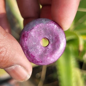 Può includere: Una gemma rotonda e viola con un foro al centro. La pietra è tenuta tra le dita su uno sfondo di fogliame verde. La gemma ha una superficie lucida con variazioni di colore. Il diametro è di circa 3 cm.