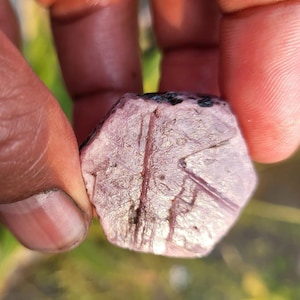 May include: A rough-cut, hexagonal-shaped gemstone with a pinkish-purple hue. The stone has visible surface lines and dark inclusions. The gem is held between fingers, with a blurred green background.