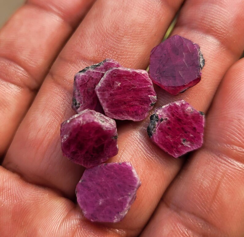 May include: A collection of raw ruby gemstones, each with a deep red hue and rough, natural edges. The gemstones are held in a hand, showcasing their natural beauty and potential for jewelry making or collecting.