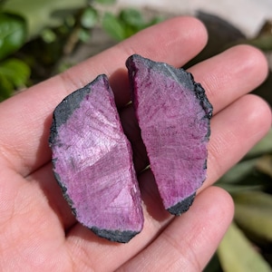Op de afbeelding: Twee ruwe, ongepolijste edelsteenschijven met een levendig magenta-roze centrum en een natuurlijke, donkere, getextureerde zwarte rand. De oppervlakken van deze stenen vertonen een kristallijne, licht reflecterende textuur met strepen.
