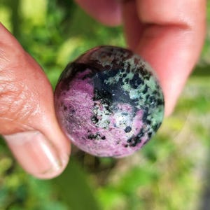 Könnte beinhalten: Eine polierte Edelstein-Kugel mit einer Mischung aus Farben. Die Kugel hat eine Basis aus Rosa und Schwarz, mit Flecken in Grün und Schwarz. Der Edelstein wird zwischen zwei Fingern vor einem verschwommenen grünen Hintergrund gehalten.