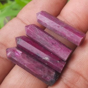 Może przedstawiać: Wyświetlane są cztery fasetowane, głęboko magentowe różdżki rubinu. Każda różdżka ma sześciokątny kształt i wypolerowaną, gładką powierzchnię. Różdżki są ułożone po przekątnej, prezentując swój bogaty kolor i krystaliczną strukturę.