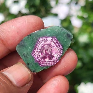 Könnte beinhalten: Ein roher, flacher Edelsteinschliff mit einem leuchtend magentafarbenen hexagonalen Kern, der von einer texturierten, undurchsichtigen grünen Außenschicht umgeben ist. Der Stein zeigt ein ausgeprägtes geometrisches Muster mit natürlichen Einschlüssen.