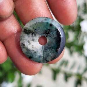 May include: A circular stone pendant with a central hole, featuring a mottled pattern of white, deep green, and dark charcoal gray mineral inclusions throughout the polished surface, creating a unique natural appearance in this stone disc.
