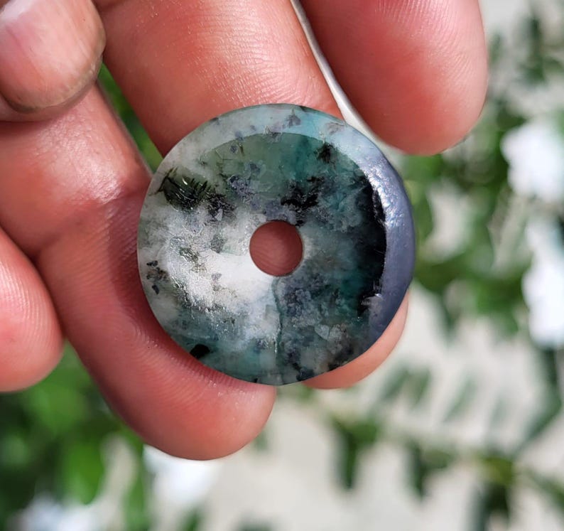 May include: A circular stone pendant with a central hole, featuring a mottled pattern of white, deep green, and dark charcoal gray mineral inclusions throughout the polished surface, creating a unique natural appearance in this stone disc.