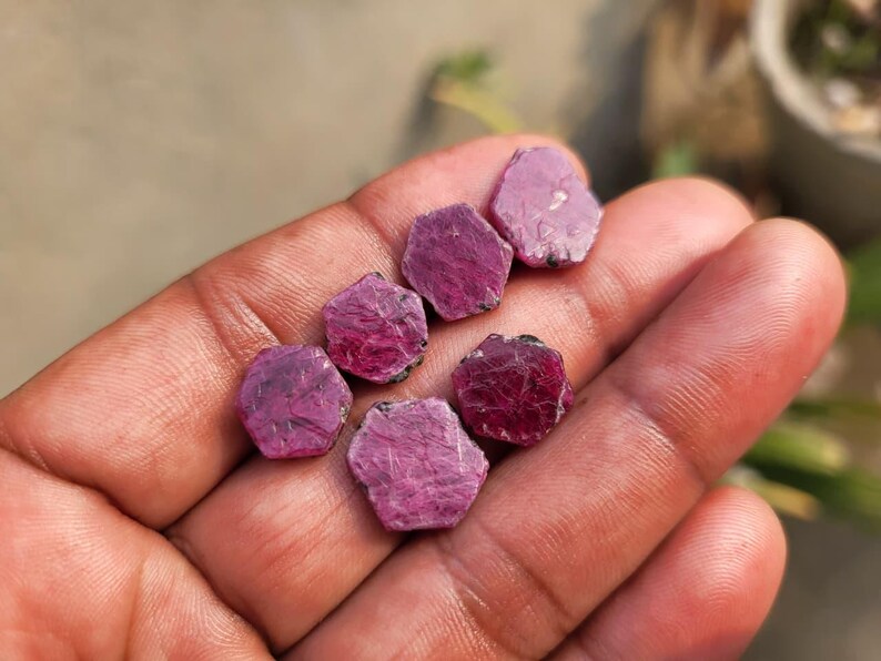 May include: A hand holds several rough-cut ruby gemstones. The gems are a deep pinkish-red color, with a slightly textured surface. The stones are irregularly shaped, with a hexagonal appearance. The background is blurred.