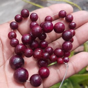 May include: A strand of deep red, round beads, varying in size, strung together on a thin, clear cord. The beads have a polished, glossy finish and are held in a person's hand, suggesting they are ready to be worn as jewelry.