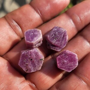 Könnte beinhalten: Vier grob geschnittene, sechseckige Rubin-Edelsteine in verschiedenen Rosa- und Lilatönen werden in einer offenen Hand gehalten. Die Edelsteine haben ein natürliches, unpoliertes Aussehen mit sichtbaren Texturen und Einschlüssen. Die Hand ist unscharf, wobei die Edelsteine im Mittelpunkt stehen.
