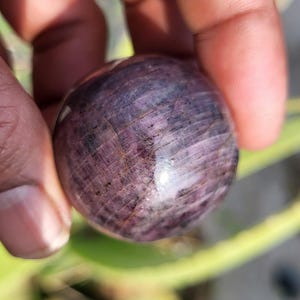 May include: A polished, spherical gemstone with a deep purple hue, featuring subtle internal veining and a glossy surface. The stone is held in a hand, showcasing its smooth, rounded form and natural beauty. The gemstone's colour ranges from dark to light purple.