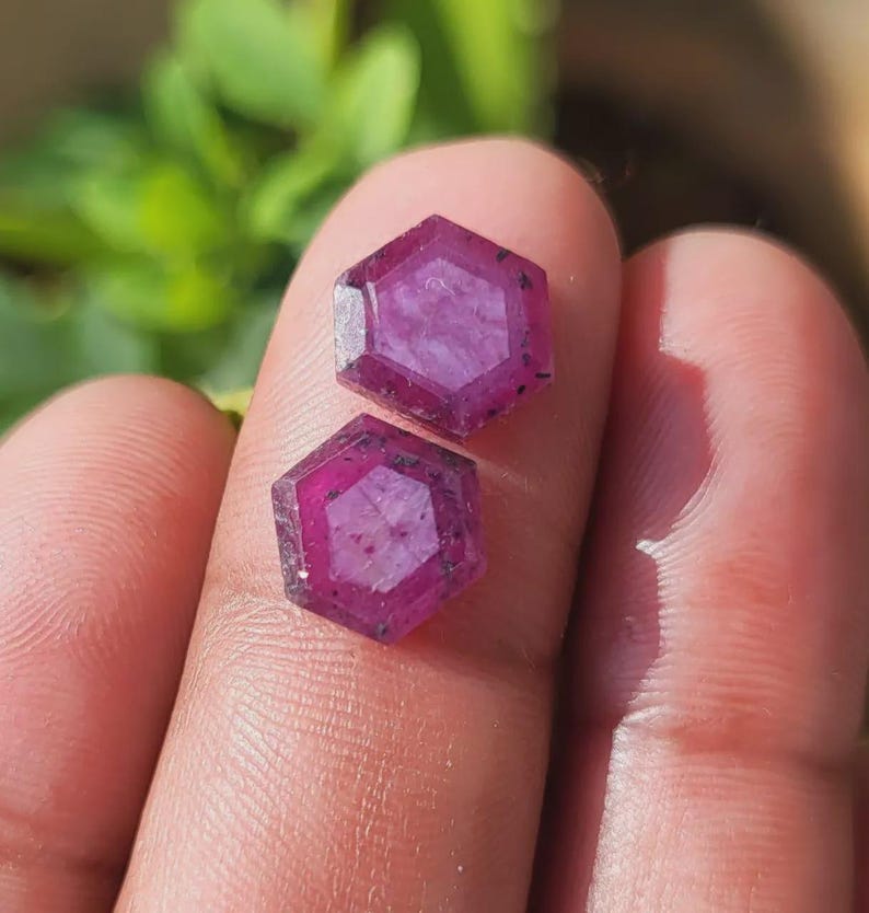 May include: Two hexagonal, faceted gemstones with a deep magenta hue are displayed. The gems have a multi-faceted design and are held between fingers. The background is blurred, with green foliage visible.