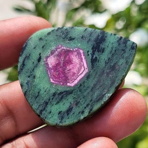 May include: A polished, teardrop-shaped zoisite gemstone featuring a vibrant, hexagonal magenta ruby crystal inclusion at its centre. The stone displays a mottled green and black matrix with a smooth, flat surface and rounded edges.