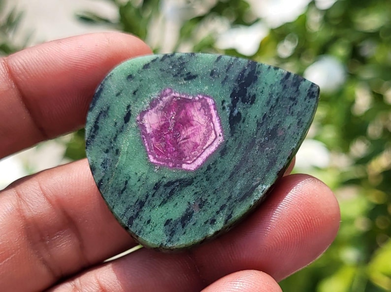 May include: A polished, teardrop-shaped zoisite gemstone featuring a vibrant, hexagonal magenta ruby crystal inclusion at its centre. The stone displays a mottled green and black matrix with a smooth, flat surface and rounded edges.