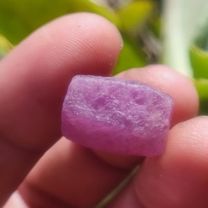 May include: A close-up of a rough, rectangular, pink gemstone. The stone has a textured surface and is held between fingers. The background is blurred green and yellow.