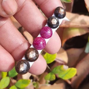 May include: A collection of faceted gemstones in shades of brown and pink are displayed on a white strip. The gemstones are round and have a sparkling appearance. The background is blurred, with green leaves and brown foliage.