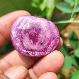 May include: A polished, oval gemstone in a deep magenta colour. The stone displays a crystalline structure with visible internal patterns and inclusions. The gem is held in a hand, with green foliage in the background.