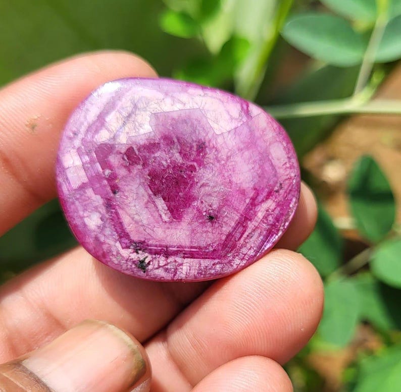 May include: A polished, oval gemstone in a deep magenta colour. The stone displays a crystalline structure with visible internal patterns and inclusions. The gem is held in a hand, with green foliage in the background.
