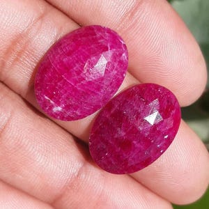 May include: Two oval-shaped, faceted rubies. The gemstones are a deep, vibrant red color with a polished surface. The rubies are held in a hand, showcasing their size and color.