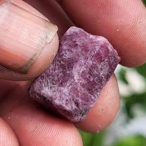 May include: A raw, unpolished gemstone with a deep magenta and purple hue. The stone features a rough, crystalline texture with visible internal fractures and natural facets across its surface, displaying a translucent quality in certain areas.