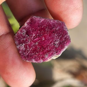 Peut inclure: Une pierre précieuse brute de rubis rouge foncé. Le rubis a une surface texturée avec des structures cristallines visibles et un bord sombre. La pierre est tenue entre deux doigts.
