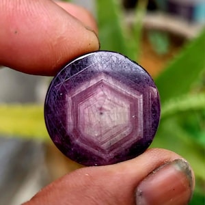 May include: A round, polished gemstone with a deep purple hue. The stone features a six-sided, star-like pattern in a lighter shade of purple at its center. Fine lines radiate from the center, creating a unique visual texture. The gemstone is held between fingers.
