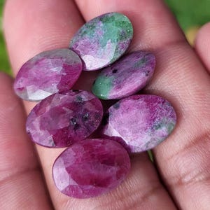 Può includere: Sei gemme ovali in varie tonalità di rosso e verde. Le pietre sono tenute in una mano, rivelando le loro superfici lucide e le variazioni naturali di colore. Le gemme sono state tagliate con delle sfaccettature per catturare la luce. Ideale per la creazione di gioielli.