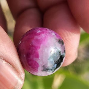 Op de afbeelding: Een gepolijste, ronde edelsteen met een mix van roze, wit en zwart. De steen wordt tussen vingers gehouden, wat het gladde, reflecterende oppervlak benadrukt. De kleuren vloeien in elkaar over, waardoor een uniek patroon ontstaat.