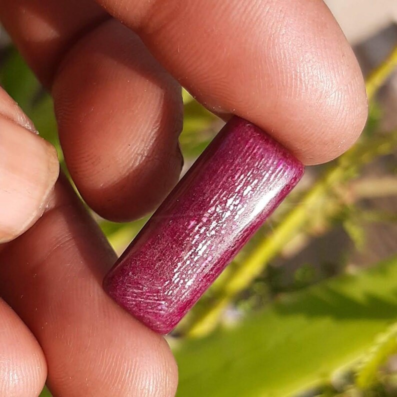 May include: A rectangular, polished gemstone in a deep burgundy hue. The stone is held between fingers, showcasing its smooth, reflective surface and subtle internal grain. The gemstone's shape is a long, slender rectangle.