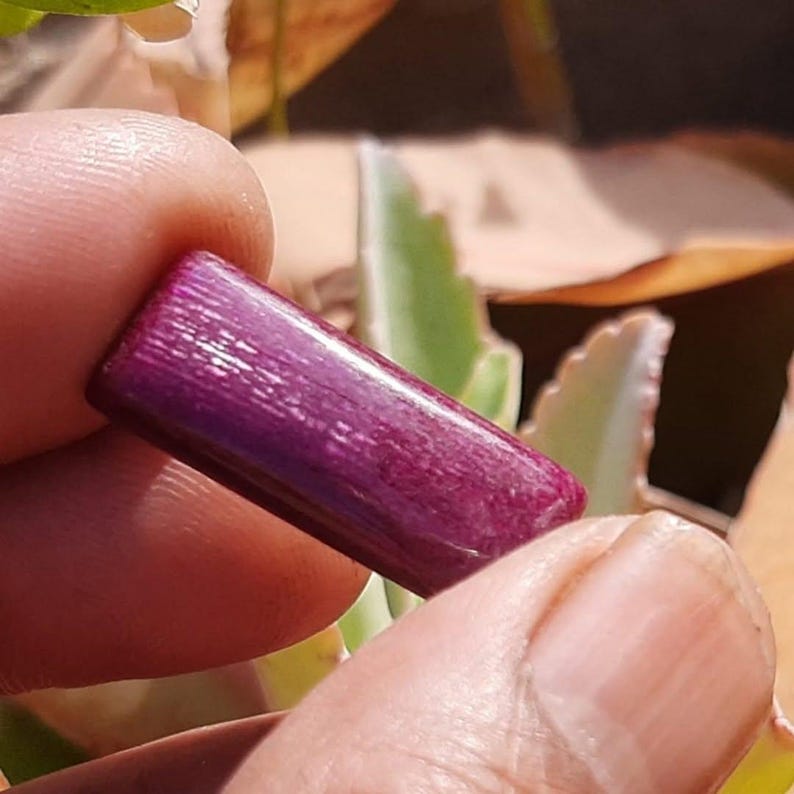 May include: A rectangular, deep purple gemstone with a textured surface. The gemstone is held between two fingers, with a blurred background of green and brown foliage.