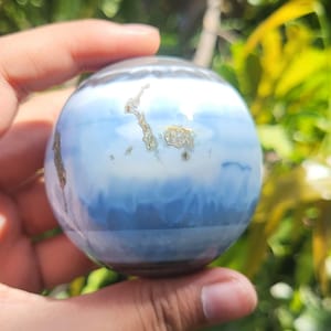 May include: A polished spherical stone featuring horizontal bands of white and various shades of blue. The surface displays a smooth, glossy texture with small, irregular, metallic-coloured inclusions scattered across the upper portion of the sphere.