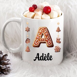Peut inclure: Mug en céramique blanche rempli de guimauves, de baies rouges et de cannelle. La tasse présente la lettre "A" en pain d'épices et le nom "Adèle". Des ornements de Noël décoratifs sont suspendus en haut de la tasse.